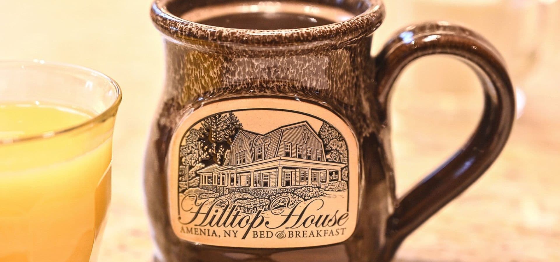 A coffee mug featuring an illustration of the Hilltop House bed and breakfast, beside a glass of orange juice.