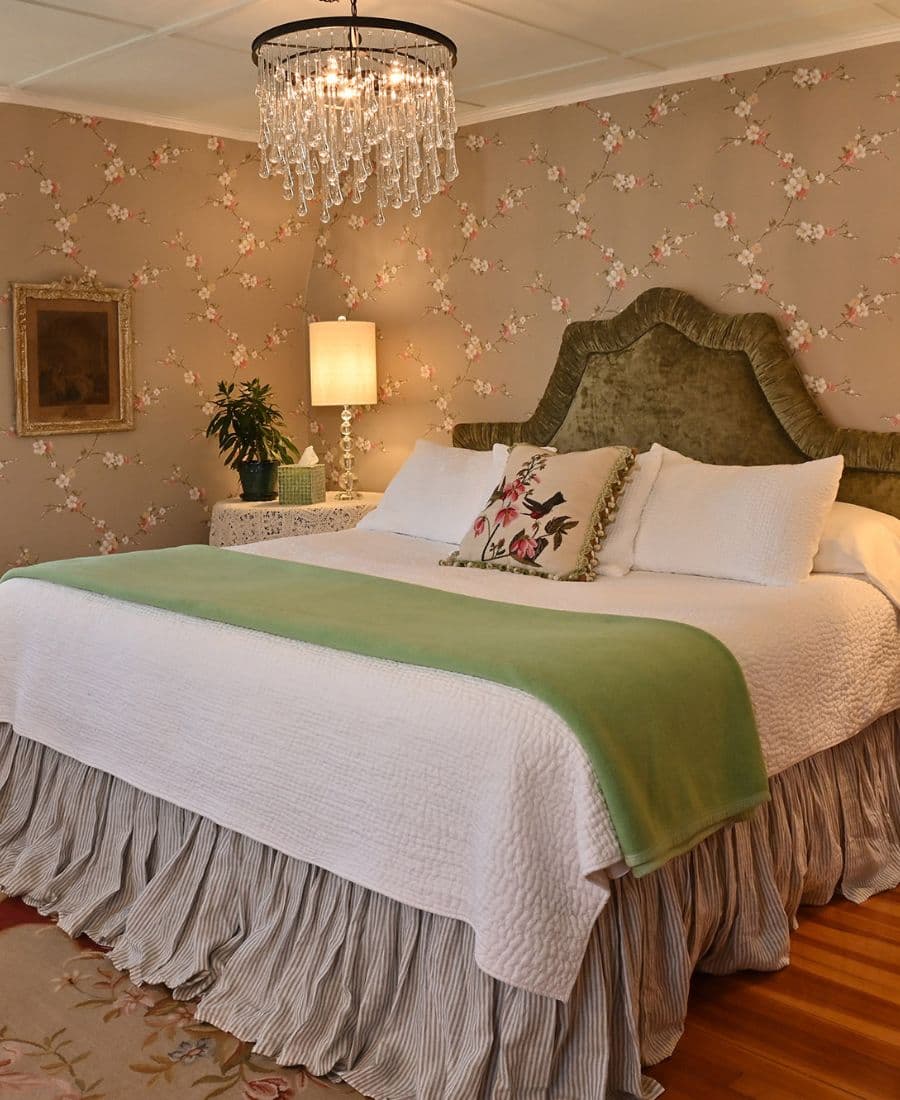 A cozy bedroom with a green-accented bed, floral wallpaper, and a crystal chandelier.