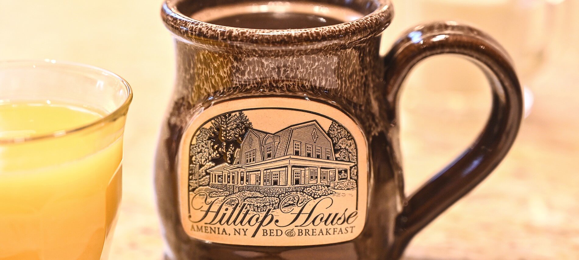 Brown pottery mug with logo of home and text Hilltop House Bed & Breakfast next to glass of orange juice